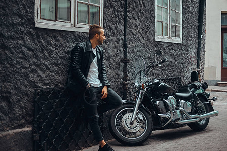 穿着黑色皮夹克和牛仔裤的时尚骑自行车的人靠在欧洲老街上他的复古摩托图片