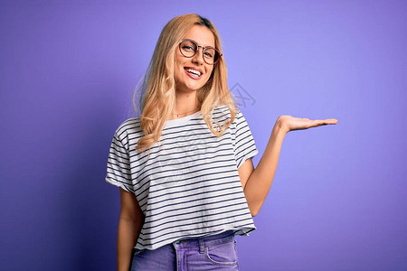 身穿条纹T恤和眼镜的金发年轻美女穿着紫色背景笑地展示并用手掌指着图片