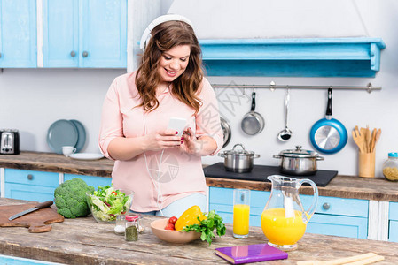 手持耳机在餐桌上用智能手机和家里厨房新鲜蔬菜的图片