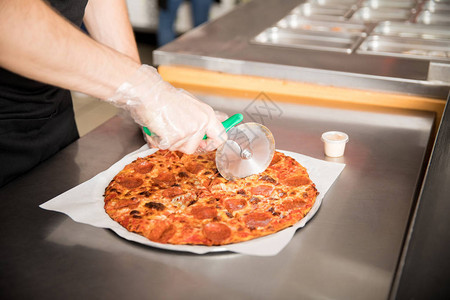 比萨店厨房柜台上戴一次手套用滚刀切比萨片的图片