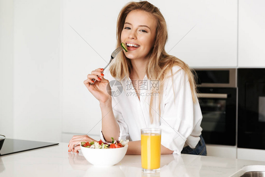 可爱的年轻女子在厨房里站着吃沙拉喝橘子汁图片