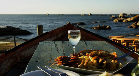 白葡萄酒和一盘海鲜在日落时在海滩上一家咖啡馆的旧船桌边有图片