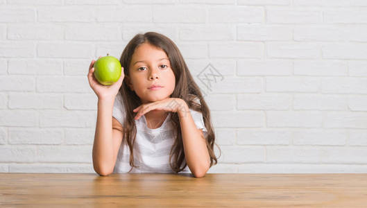 坐在桌子上吃新鲜绿苹果的年轻西班牙小孩在聪明的面孔上自信地表情图片