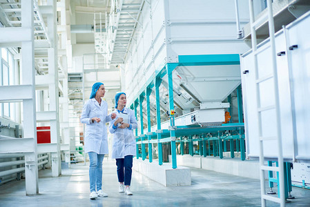 两名身穿实验室大衣的年轻女工在清洁生产车间复制空间中通过电力单位横跨大厅行走的完整背景图片