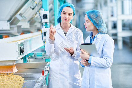 清洁生产车间两名身穿实验室外套的年轻女工站在带通心粉的输图片