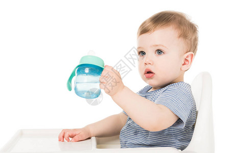 可爱的小可爱男孩拿着带水的婴儿杯子坐在高椅子上孤背景图片