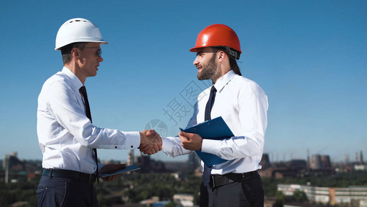 身穿硬衣站立讨论和握手的商人在背景中与工厂进行交易图片