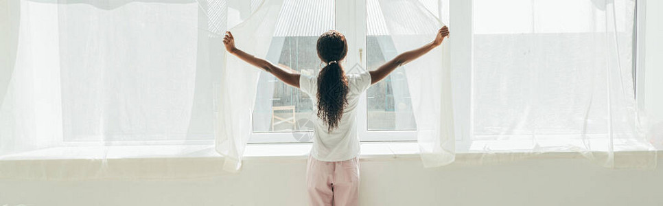 穿着睡衣的非洲裔美国女孩在阳光下打开窗帘的后图片