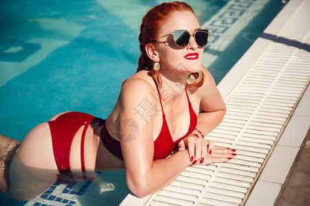 享受晒黑和假期漂亮的年轻女子穿着红色泳装躺在游泳池附图片