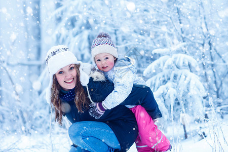 快乐的年轻妈和女儿在雪花背景雪林中的冬帽中图片