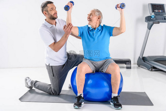 康复治疗师协助老年男子在健身球上用哑铃进图片