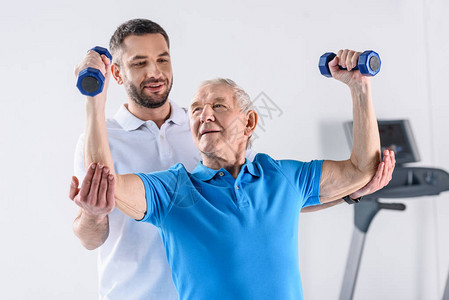 协助老年男子在灰色背景下用哑铃进行锻炼的康图片