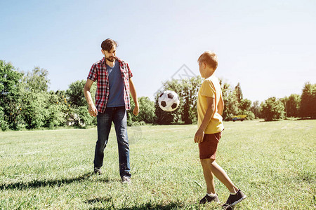 父亲和儿子踢足球比赛的照片有一个球从爸传给儿子它在空气中田野图片