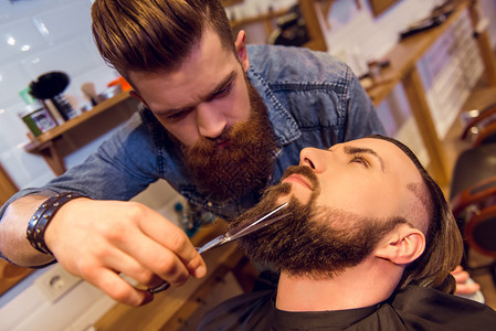 在理发店剪有吸引力的客户胡须的英俊年轻胡子图片