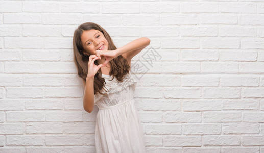 在白砖墙上笑着爱的年轻西班牙小孩展示了心脏符号和双手的形状图片