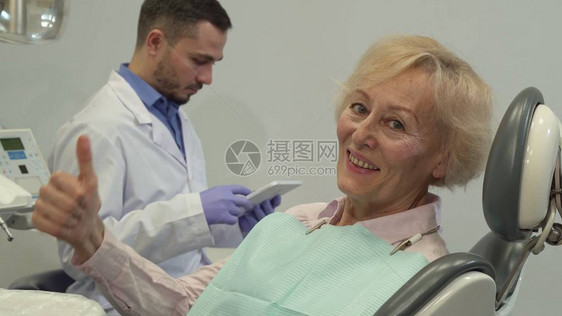 高级女客户在牙科椅上竖起大拇指有魅力的白种女人批准看牙医的预约相机沿着金发碧眼的老妇人向上倾斜图片