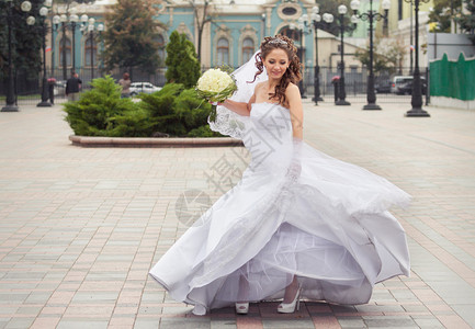 穿着白色婚纱的美丽黑发新娘拿着白玫瑰花朵和在城市公园背景上跳背景图片