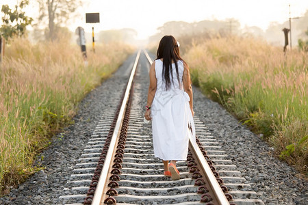 年轻女子走出摄像头走在一条农村铁路的铁轨上在夏天晚上穿着白色衣服长图片