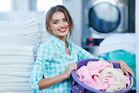 女人拿着篮子和玫瑰色亚麻布在洗衣店感觉亚麻布的柔软和洗涤的新鲜感女孩靠近洗衣机和成堆的白色亚麻背景图片