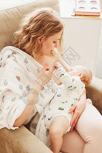 年轻美丽的中年金发白人母亲抱着母乳喂养婴儿坐在扶手椅上的肖像图片