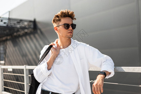 穿着白色T恤戴着时髦太阳镜穿着时髦衬衫酷的年轻潮人站在城市街道上的金属栅栏附近现代男模特喜欢在阳光明图片