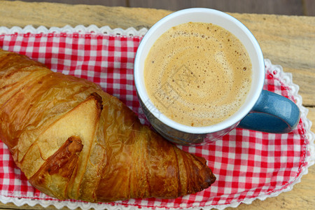 蓝色咖啡杯与新鲜咖啡和奶油和羊角面包与红色白桌巾毛与刺绣在复古风格的旧木制早餐桌上早上图片