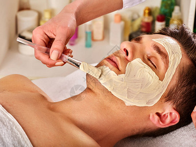 人在温泉沙龙的泥面膜用粘土全脸进行治疗按摩男躺水疗床美容师用化妆刷治疗程序我们图片