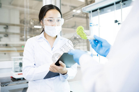 微生物学家拿着绿色食品营养测试样品的手钳和看着它的女人把结果写进剪贴板图片