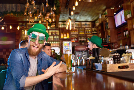 戴着滑稽派对眼镜和绿色圆顶礼帽的快乐红发男子坐在吧台前图片