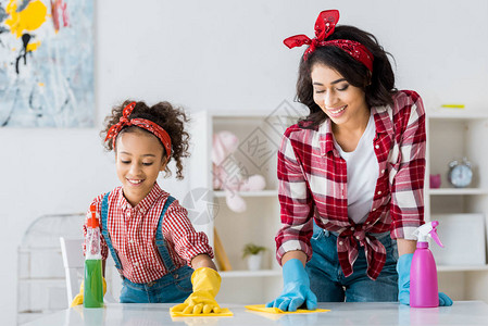 快乐的非裔美国母亲可爱的女儿清洁桌戴着明图片