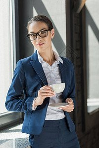 身穿西装拿着咖啡杯和看办公室摄像机的美丽自信女商人背景图片