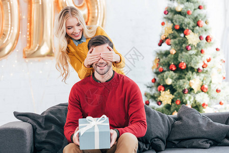 女孩闭着眼睛用礼物盒给家里有圣诞树的男朋友送礼物盒图片