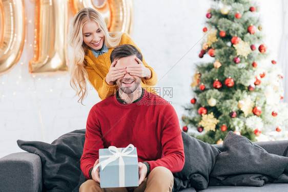 女孩闭着眼睛用礼物盒给家里有圣诞树的男朋友送礼物盒图片