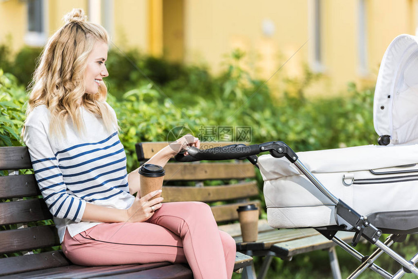 一边是微笑的母亲坐在长凳上喝着咖啡去公园里把婴儿图片