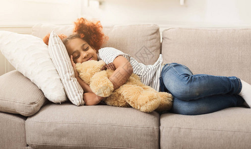 快乐的黑人小女孩在家里的沙发上睡着玩具微笑的梦幻般的孩子在沙发上打图片