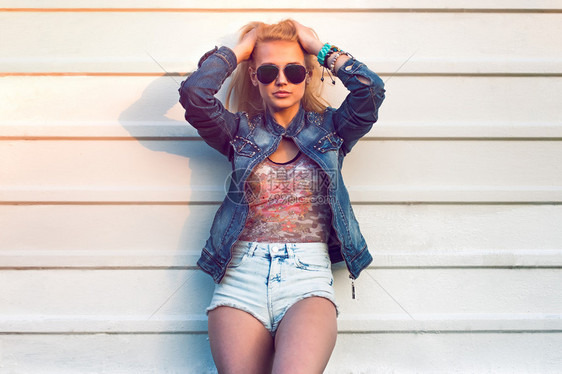 年轻时尚迷人的女人或女孩的户外夏季特写肖像在阳光明媚的日子里穿着街头牛仔裤夹克和镜站在白墙附近采用的复古色图片