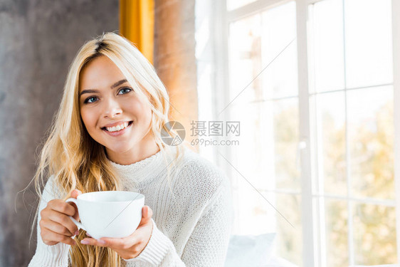 穿着毛衣的快乐美丽的女人拿着一杯咖啡早上在卧室里看摄影图片