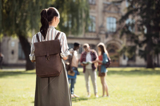 年轻女学生背着包看着同学站在大学附近的草地图片