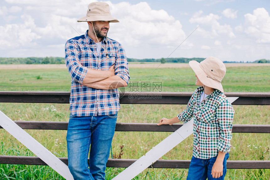 身戴巴拿马帽子的父亲和儿子在农场的栅栏附近站立时图片