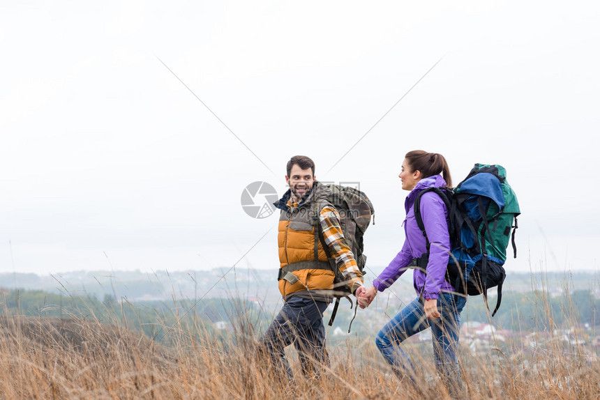 带着手牵背包和在农村地区高草中行走的年轻一对笑图片