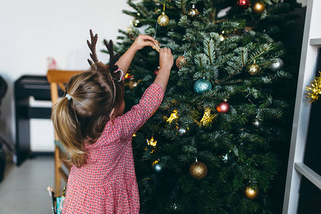 可爱的小女孩穿着有趣的节庆鹿角装饰圣诞树背景图片