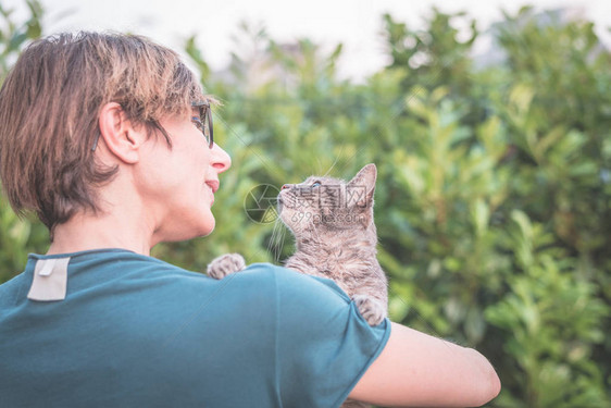 顽皮的家猫被戴眼镜的微笑女人抱着和拥抱绿色家庭花园的户外环境浅景深图片