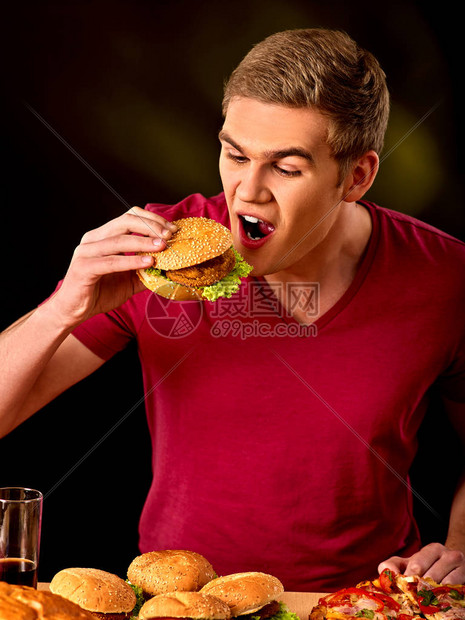 男人吃汉堡包和比萨饼学生在桌子上吃快餐的画像男试图在黑暗的背景下吃垃圾大忙图片