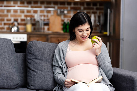 微笑的年轻孕妇在家吃苹果和看书图片