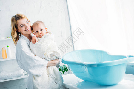 在浴袍中微笑的母亲带着可爱的孩子被毛巾覆盖在塑料婴儿浴图片