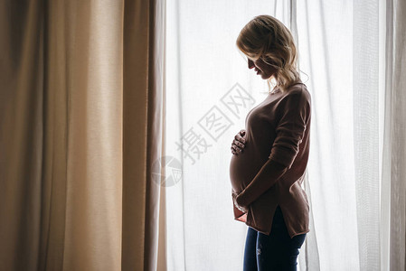 美丽的孕妇的侧面手在肚子上站在家庭窗户图片