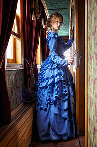 19世纪晚期穿蓝色旧礼服的年轻女子站在逆向铁路车走廊窗图片