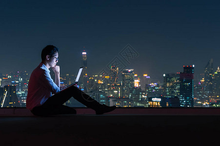 坐在大楼顶部的亚洲商人使用具有城市夜景商业成功和技术理念图片