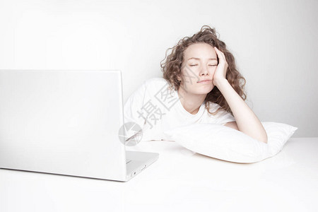 在白墙前用笔记本电脑工作时睡在工作场所的红发妇图片