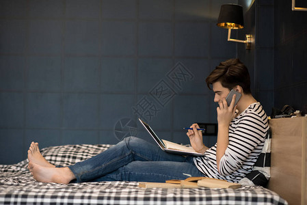青少年男孩躺在床上使用笔记本电脑和同时用电话说时做功图片